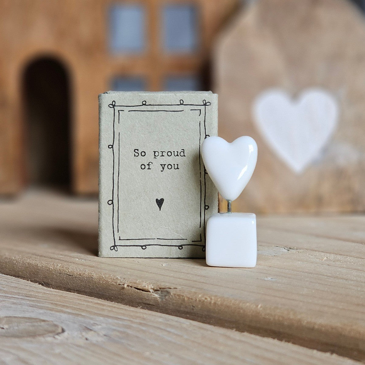 A small white ceramic star and box figurine stands beside its light grey packaging box on a wooden surface. The box is printed with the words 'So proud of you' and a black heart.