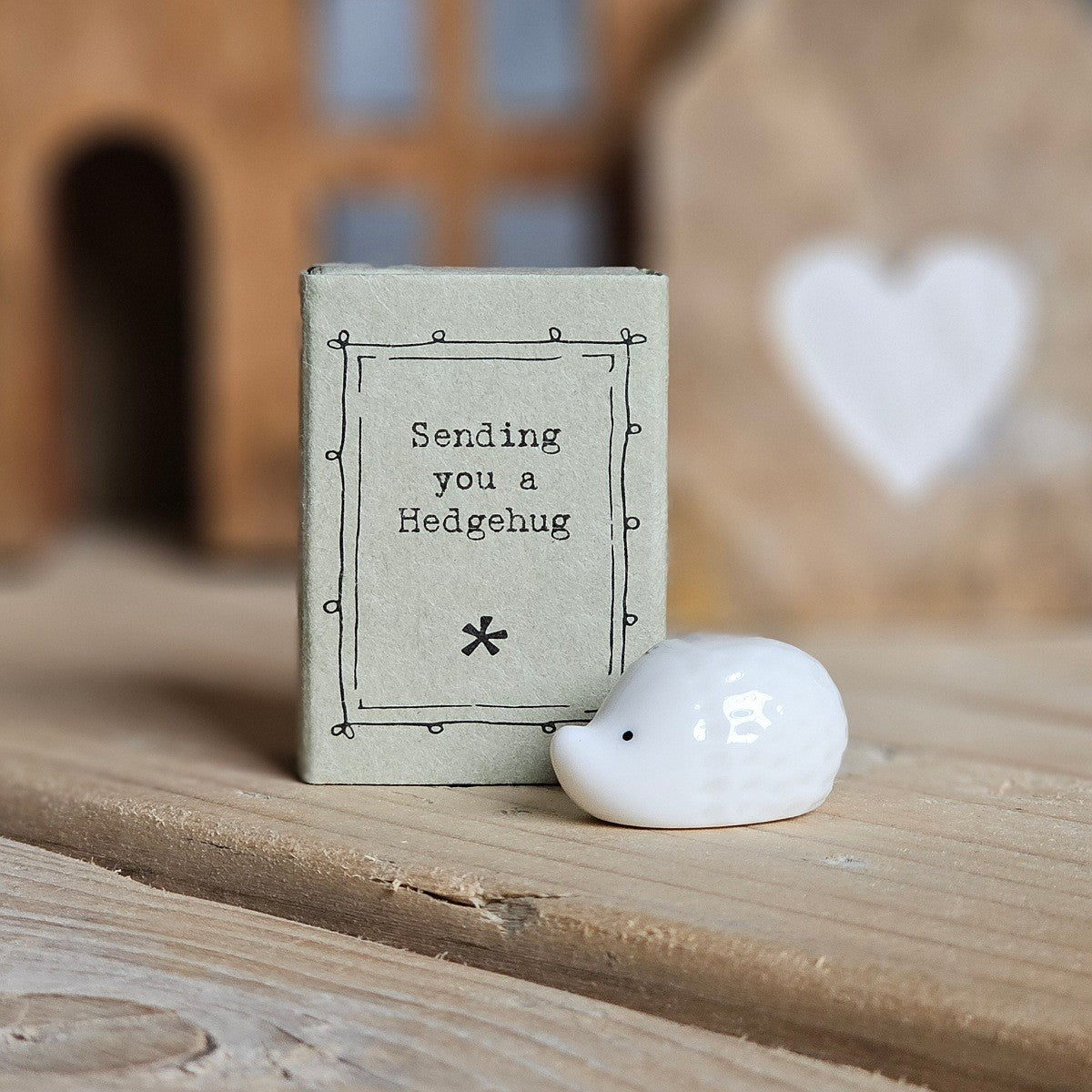 A small white ceramic hedgehog figurine stands beside its light grey packaging box on a wooden surface. The box is printed with the words 'Sending you a Hedgehug' and a black flower.