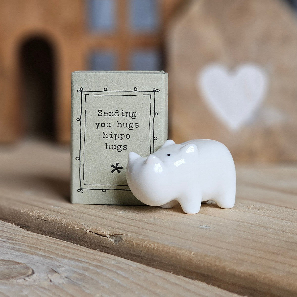 A small white ceramic hippo figurine stands beside its light grey packaging box on a wooden surface. The box is printed with the words 'Sending you huge hippo hugs' and a black flower.