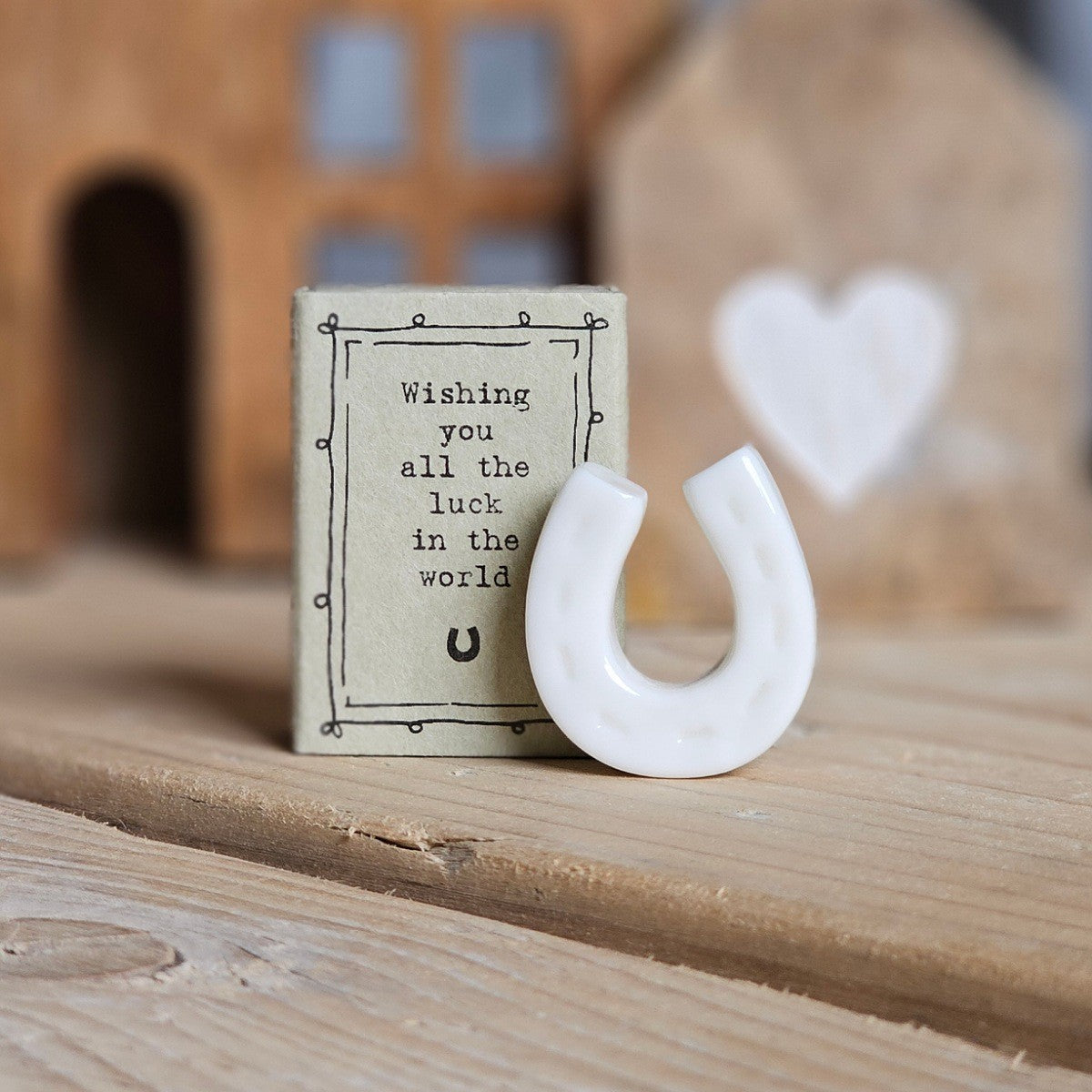 A small white ceramic horseshoe figurine stands beside its light grey packaging box on a wooden surface. The box is printed with the words 'Wishing you al the luck in the world' and a black horseshoe.