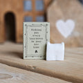 A small white ceramic house figurine stands beside its light grey packaging box on a wooden surface. The box is printed with the words 'Wishing you every happiness in your new home' and a black house.