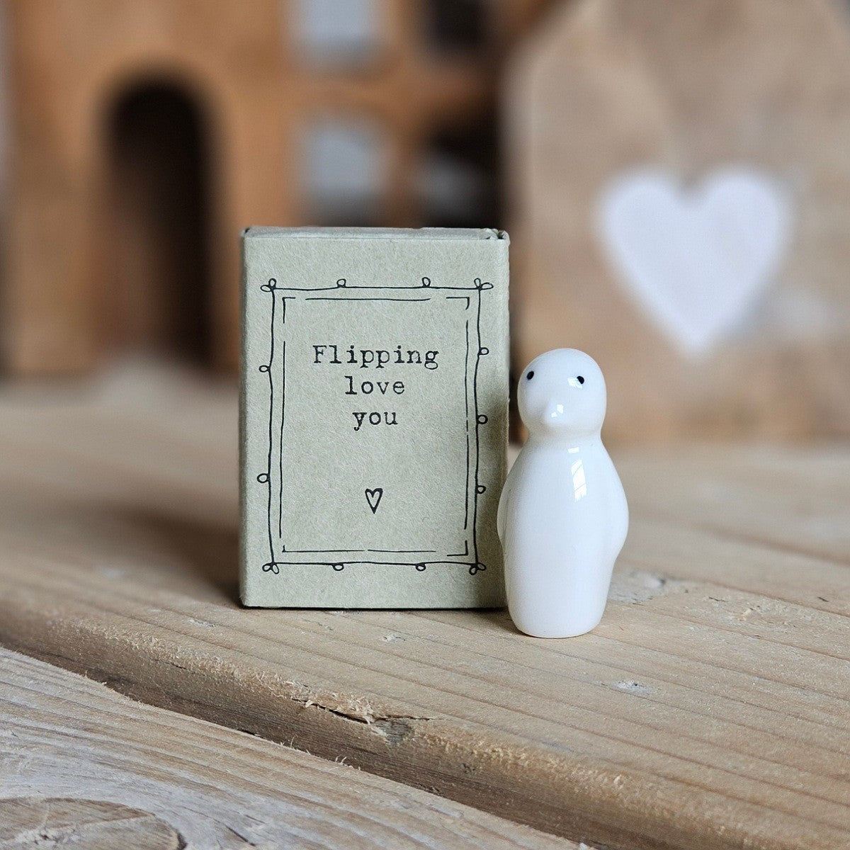 A small white ceramic penguin figurine stands beside its light grey packaging box on a wooden surface. The box is printed with the words 'Flipping love you' and a heart.