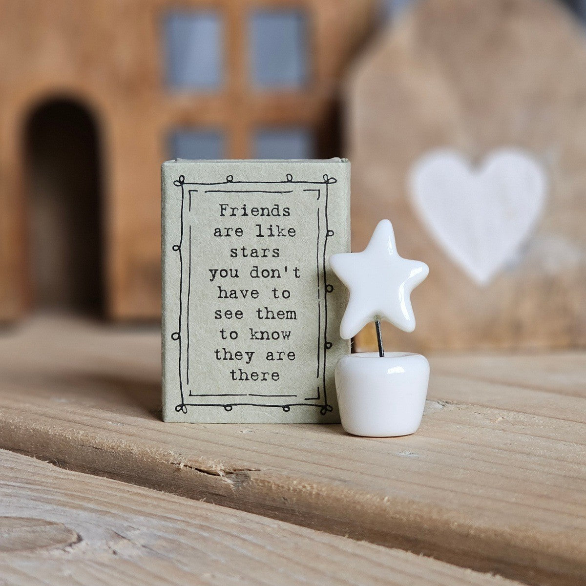 A small white ceramic star and pot figurine stands beside its light grey packaging box on a wooden surface. The box is printed with the words 'Friends are like stars you don't have to see the to know they are there'.
