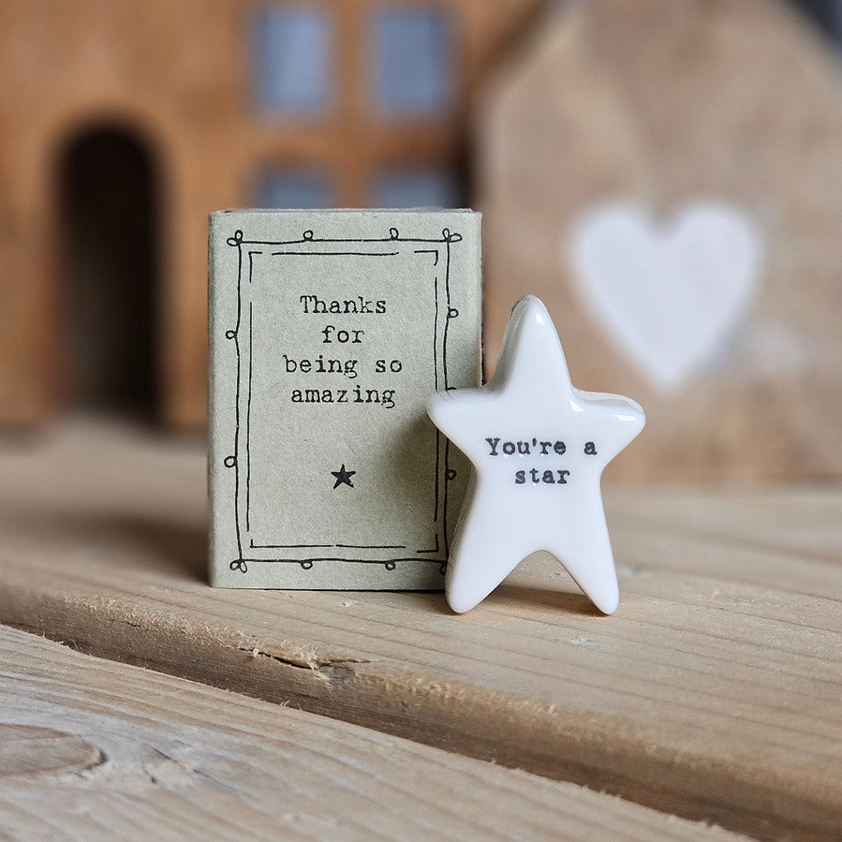 A small white ceramic star figurine with the words 'You're a star' stands beside its light grey packaging box on a wooden surface. The box is printed with the words 'Thanks for being amazing' and a black star.