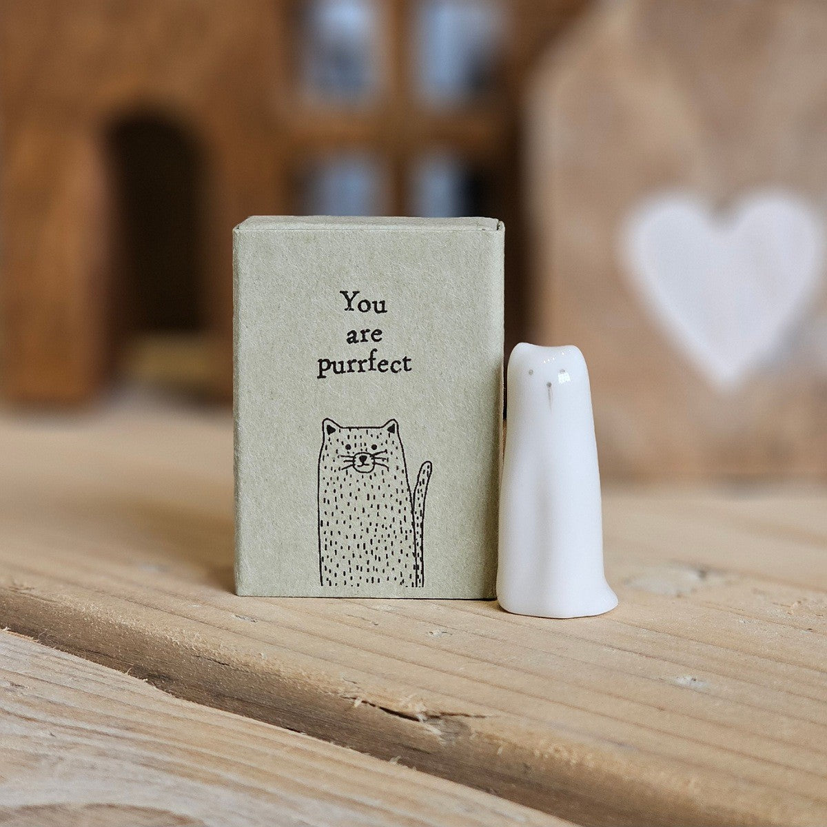 A small white ceramic tall cat figurine stands beside its light grey packaging box on a wooden surface. The box is printed with the words 'You are purrfect' and a black cat.