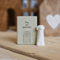 A small white ceramic tall dog figurine stands beside its light grey packaging box on a wooden surface. The box is printed with the words 'You are pawsome' and a black dog.