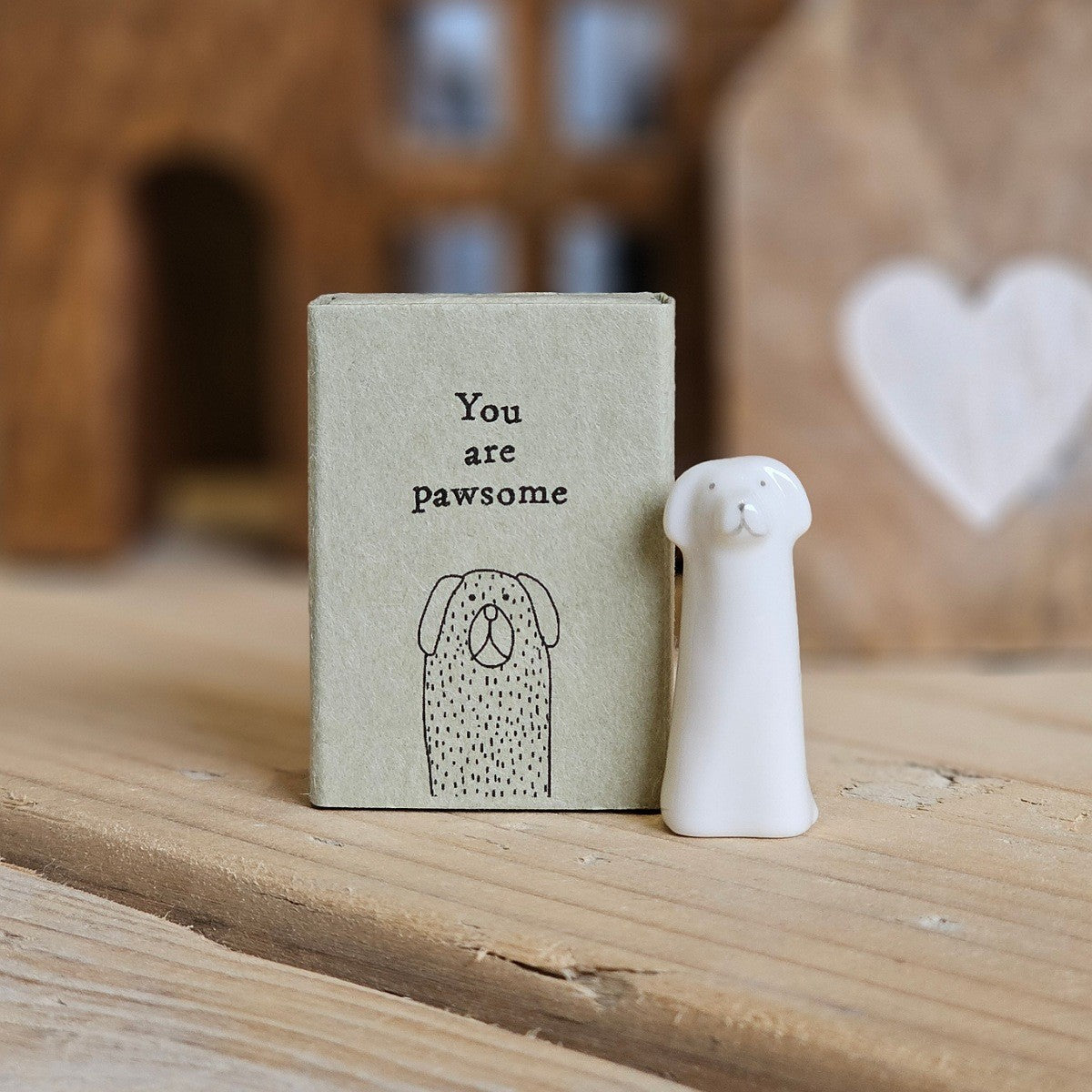 A small white ceramic tall dog figurine stands beside its light grey packaging box on a wooden surface. The box is printed with the words 'You are pawsome' and a black dog.