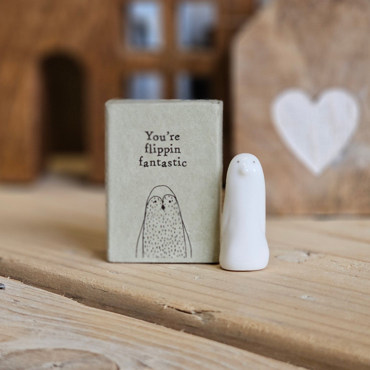 A small white ceramic penguin figurine stands beside its light grey packaging box on a wooden surface. The box is printed with the words 'You're flippin fantastic' and a black penguin.