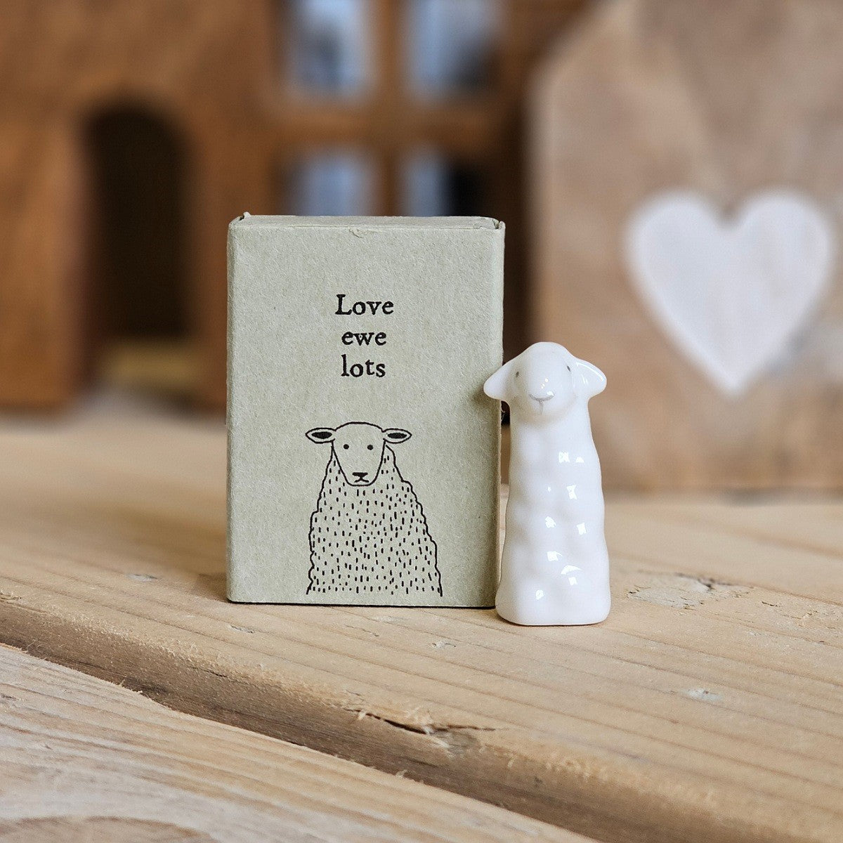 A small white ceramic sheep figurine stands beside its light grey packaging box on a wooden surface. The box is printed with the words 'Love ewe lots' and a black sheep.