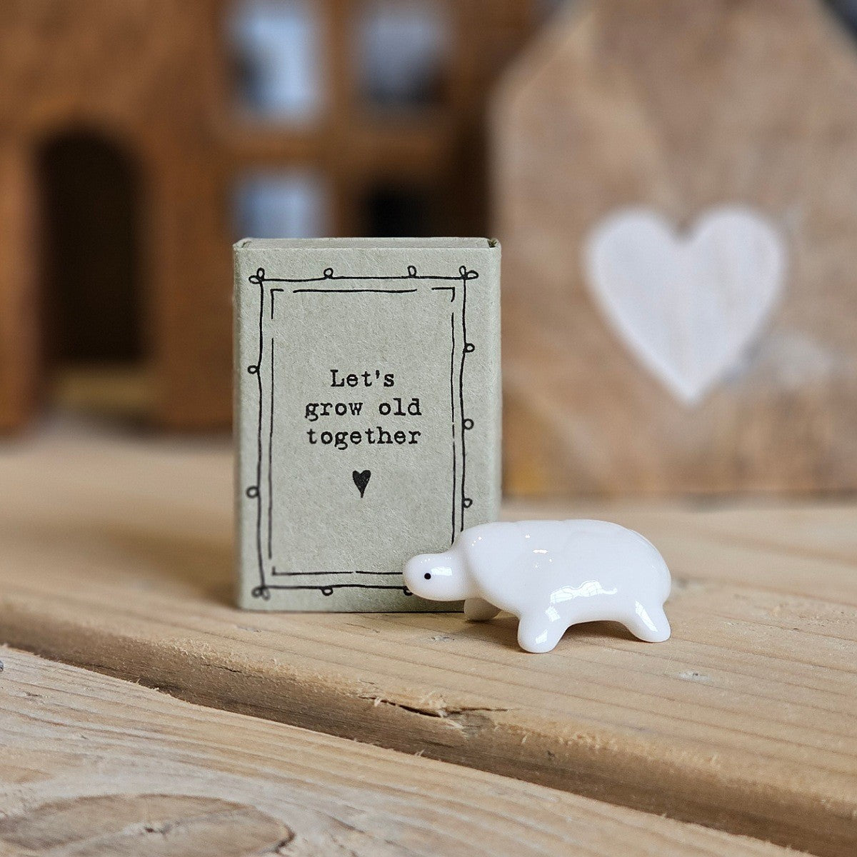 A small white ceramic tortoise figurine stands beside its light grey packaging box on a wooden surface. The box is printed with the words 'Let's grow old together' and a black heart.