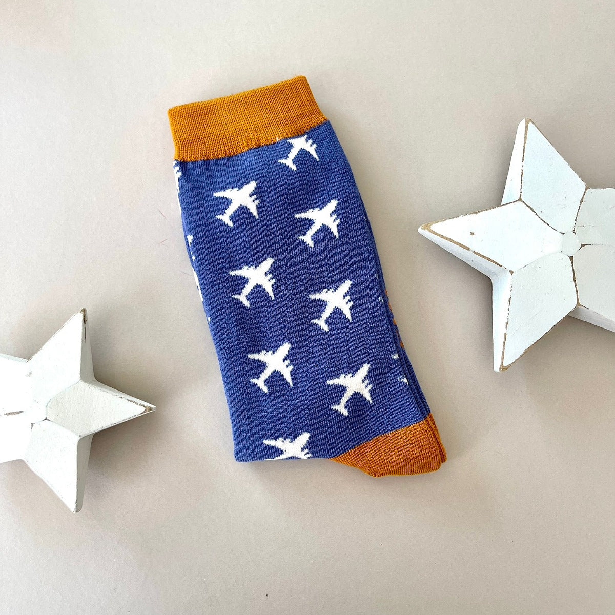 A single sock laid flat on a beige background, featuring a blue body with a pattern of white airplane print and mustard ribbed cuffs and heal. Two white star-shaped decorative props sit on either side.