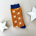 A single sock laid flat on a light beige background, featuring a mustard body with a pattern of white airplane print and navy blue ribbed cuffs and heal. Two white star-shaped decorative props sit on either side.