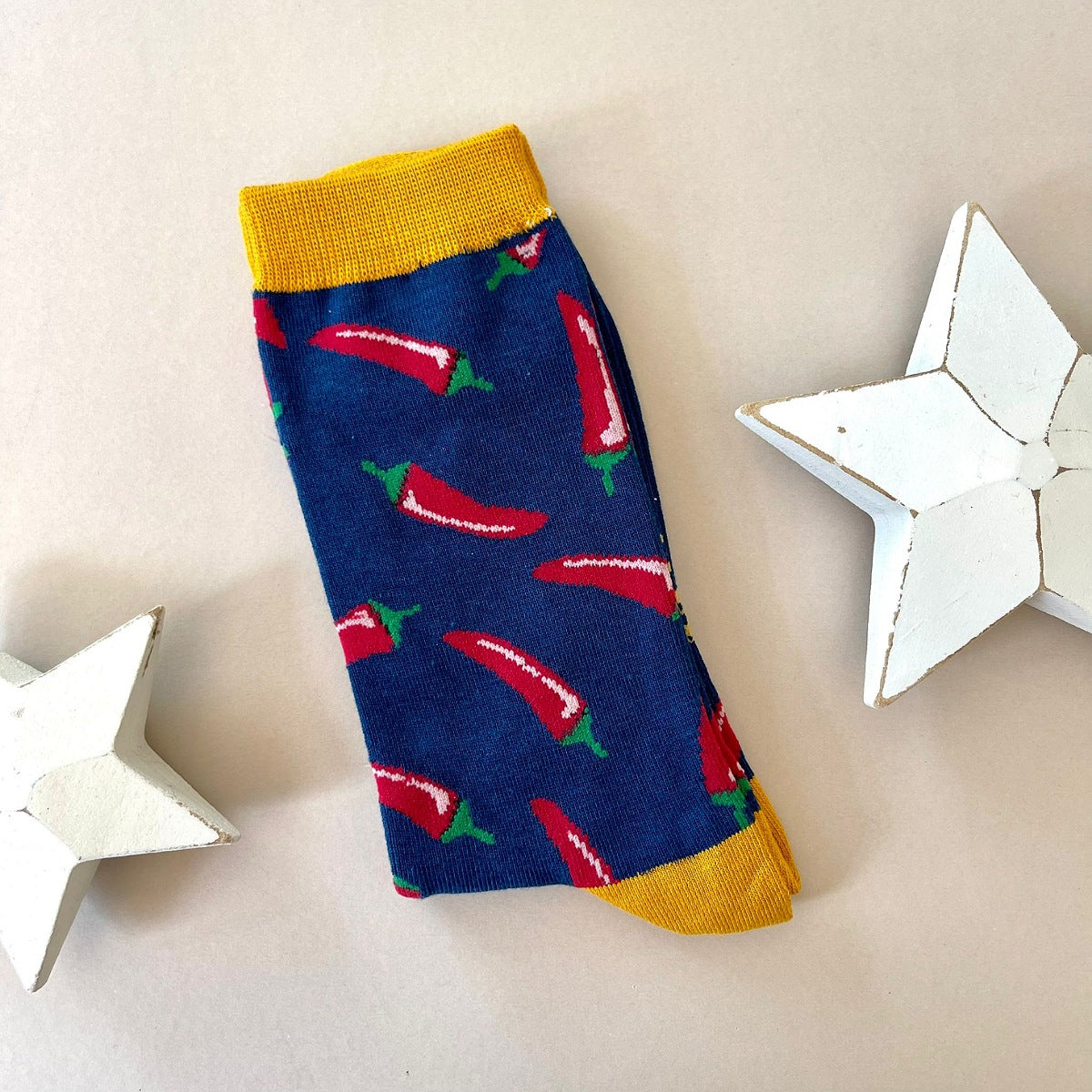A single navy blue sock with an all-over pattern of red chillis.  Mustard ribbed cuffs and heels, styled on a light beige surface with white star props.
