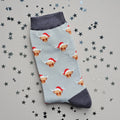 A single silver grey sock with an all-over  pattern of highland cows wearing santa hats.  Dark grey ribbed cuffs and heels.  Styled on a light grey surface with scattered silver stars props.