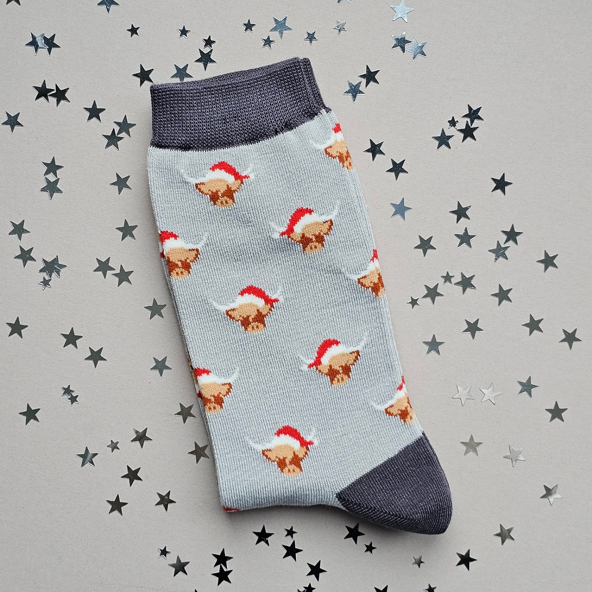 A single silver grey sock with an all-over  pattern of highland cows wearing santa hats.  Dark grey ribbed cuffs and heels.  Styled on a light grey surface with scattered silver stars props.