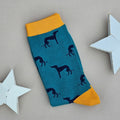 A single teal sock with an all-over pattern of navy greyhound dogs.  Yellow ribbed cuffs and heels, styled on a light grey surface with white star props.
