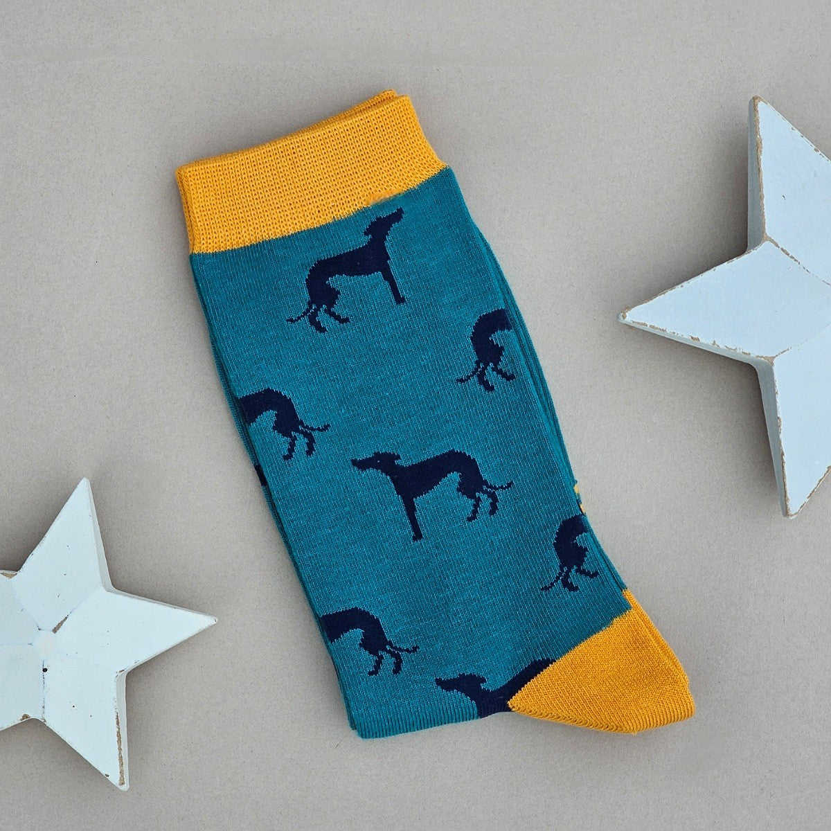 A single teal sock with an all-over pattern of navy greyhound dogs.  Yellow ribbed cuffs and heels, styled on a light grey surface with white star props.
