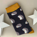 A single dark grey sock with an all-over pattern of cute hedgehogs with white face and tummy with beige spike backs.  Mustard ribbed cuffs and heels.  Styled on a light beige surface with white star props.
