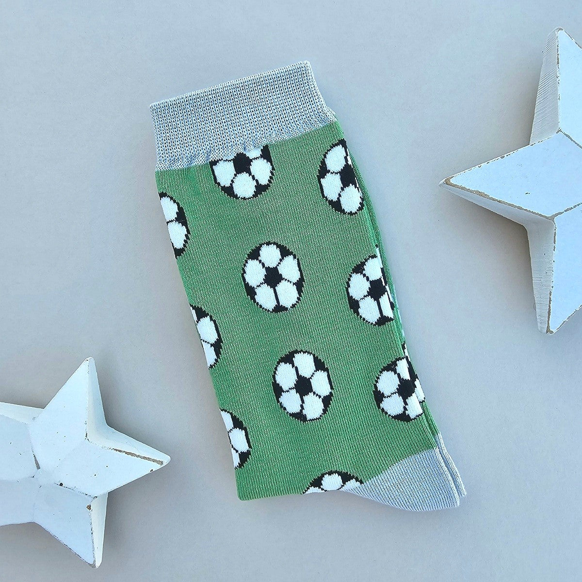 A single green sock with an all-over pattern of footballs in black and white.  silver grey ribbed cuffs and heels, styled on a light beige surface with white star props.
