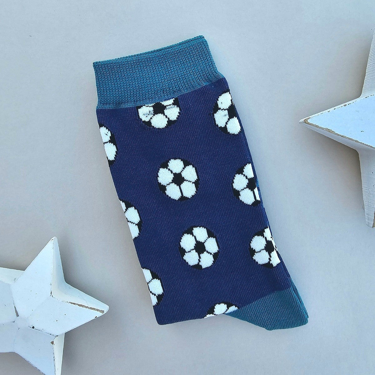A single navy blue sock with an all-over pattern of footballs in black and white.  Teal ribbed cuffs and heels, styled on a light beige surface with white star props.
