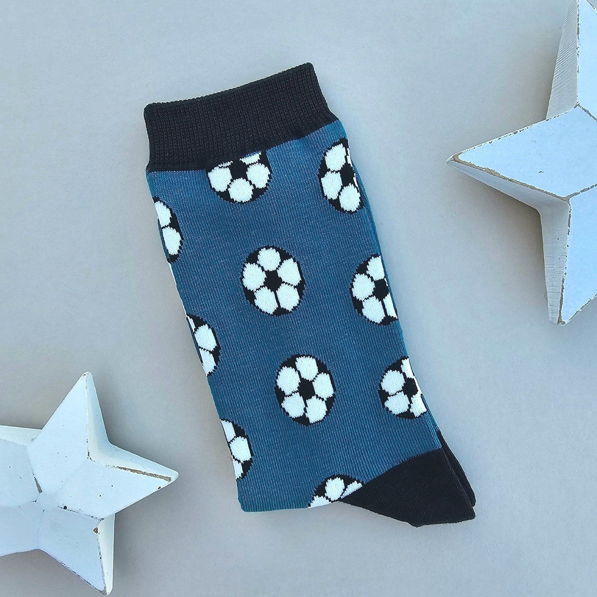 A single teal sock with an all-over pattern of footballs in black and white.  Black ribbed cuffs and heels, styled on a light beige surface with white star props.
