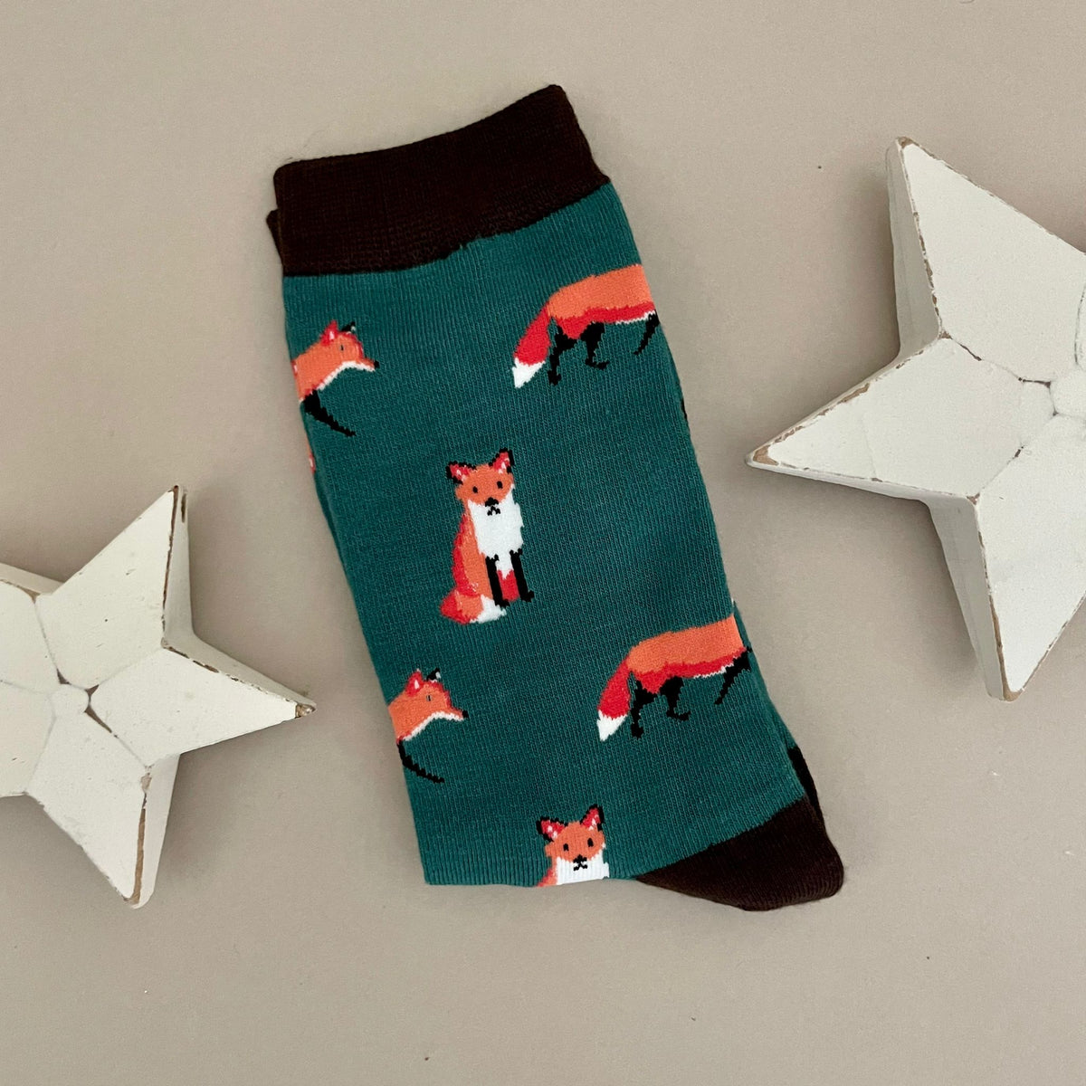 A single green sock with an all-over pattern of red and orange foxes.  Black ribbed cuffs and heels, styled on a light beige surface with white star props.
