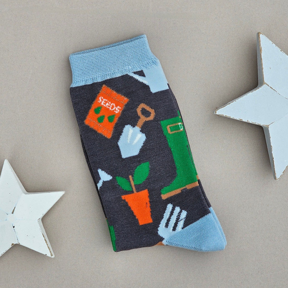 A single grey sock with an all-over  pattern of gardening items and tools in green, orange, brown and blue.  Light blue ribbed cuffs and heels, styled on a light grey surface with white star props.
