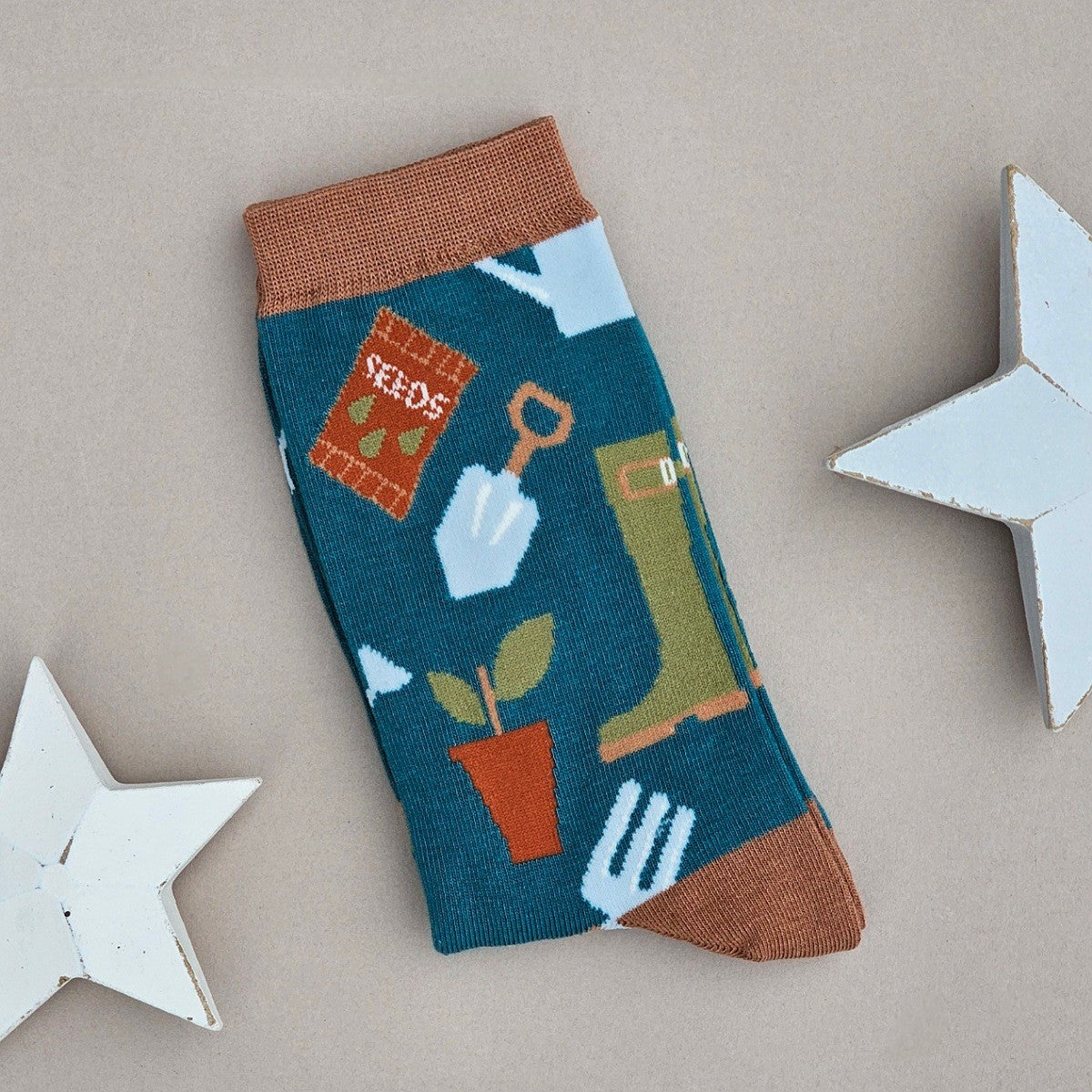 A single teal sock with an all-over  pattern of gardening items and tools in olive green, rust orange, brown and light blue.  Brown ribbed cuffs and heels, styled on a light grey surface with white star props.
