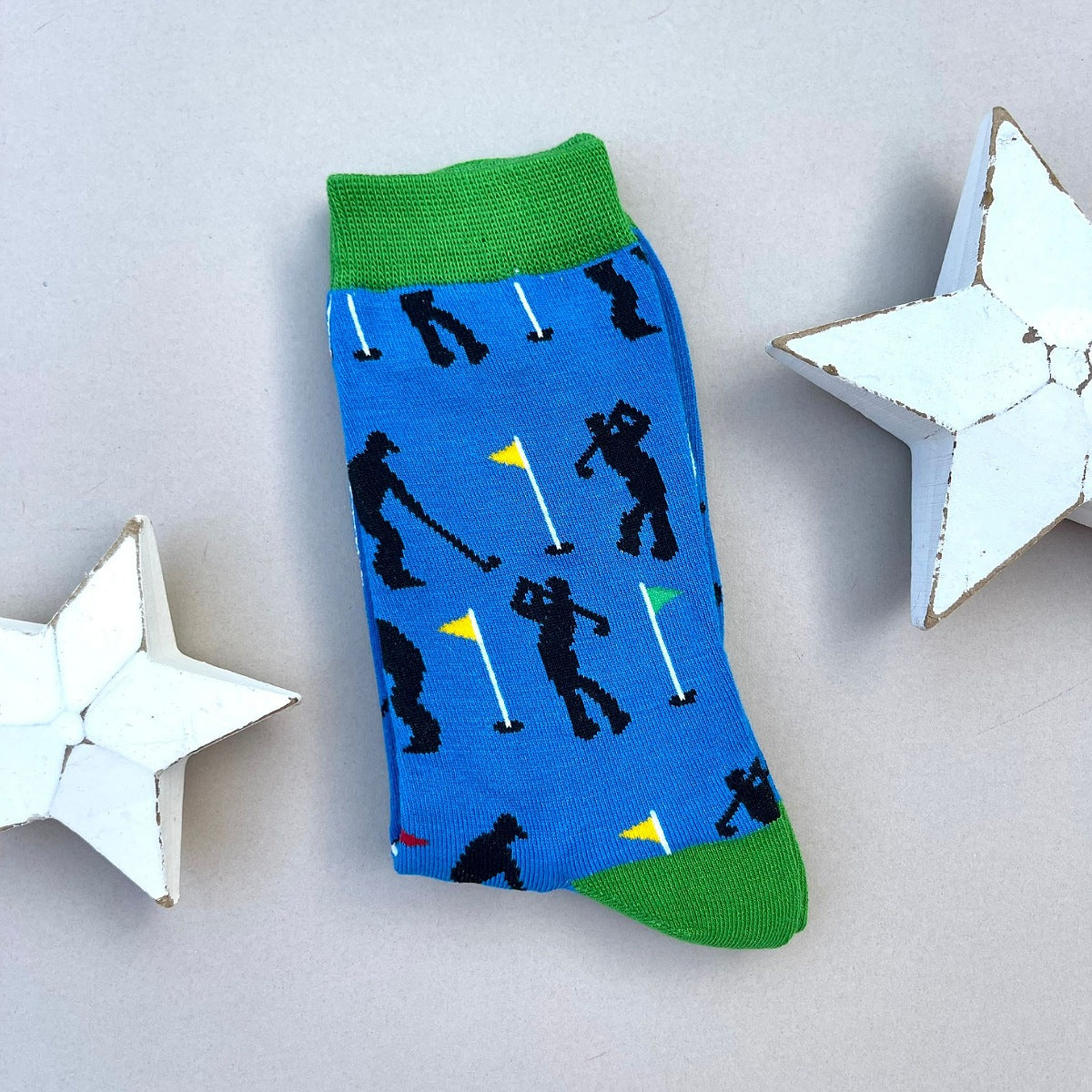 A single blue novelty sock with a green cuff and toe, featuring a pattern of black golfers and yellow/green flags, displayed on a light grey background with two white star-shaped decorative props.