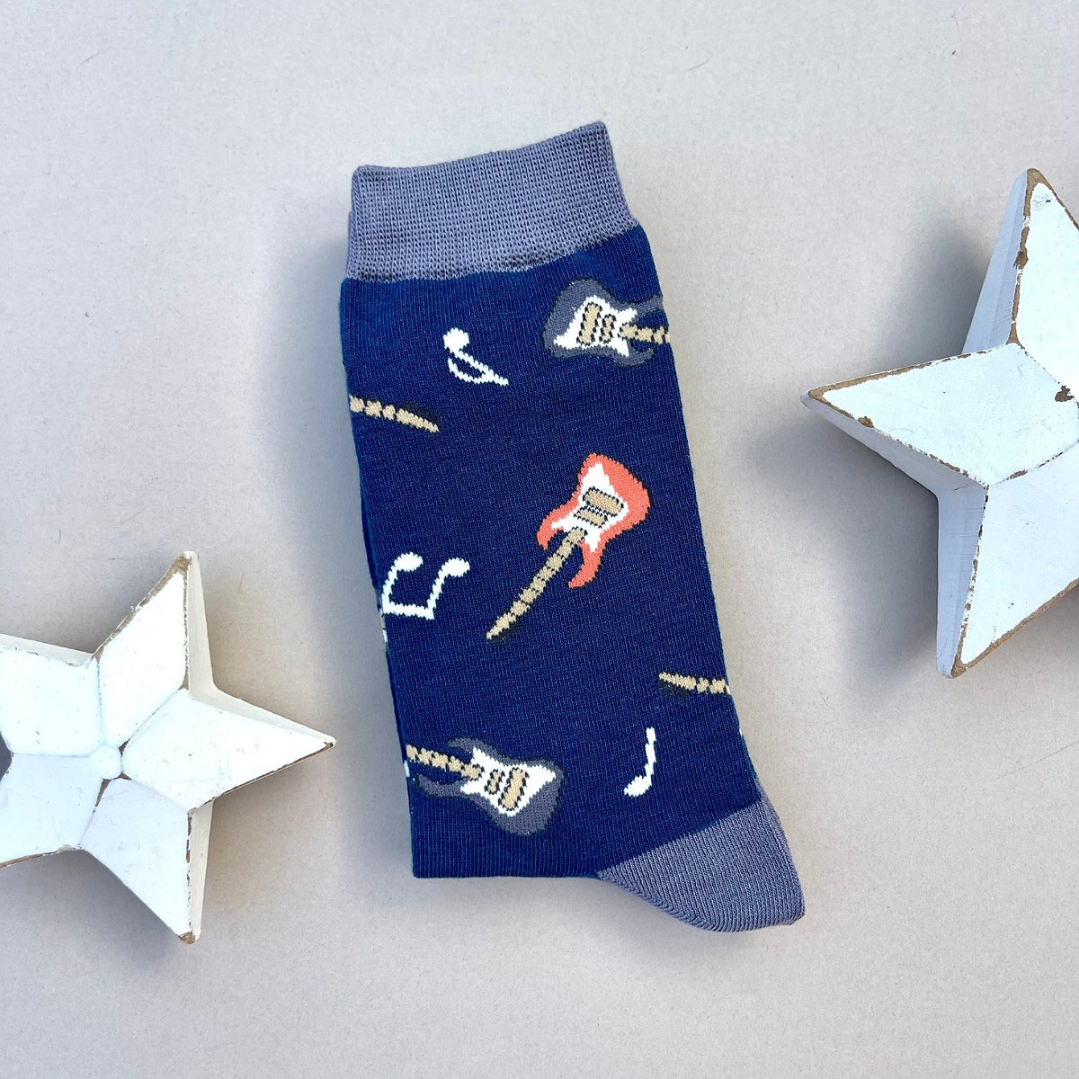 A single navy blue sock with an all-over pattern of blue and red guitars with music notes.  Blue ribbed cuffs and heels, styled on a light grey surface with white star props.
