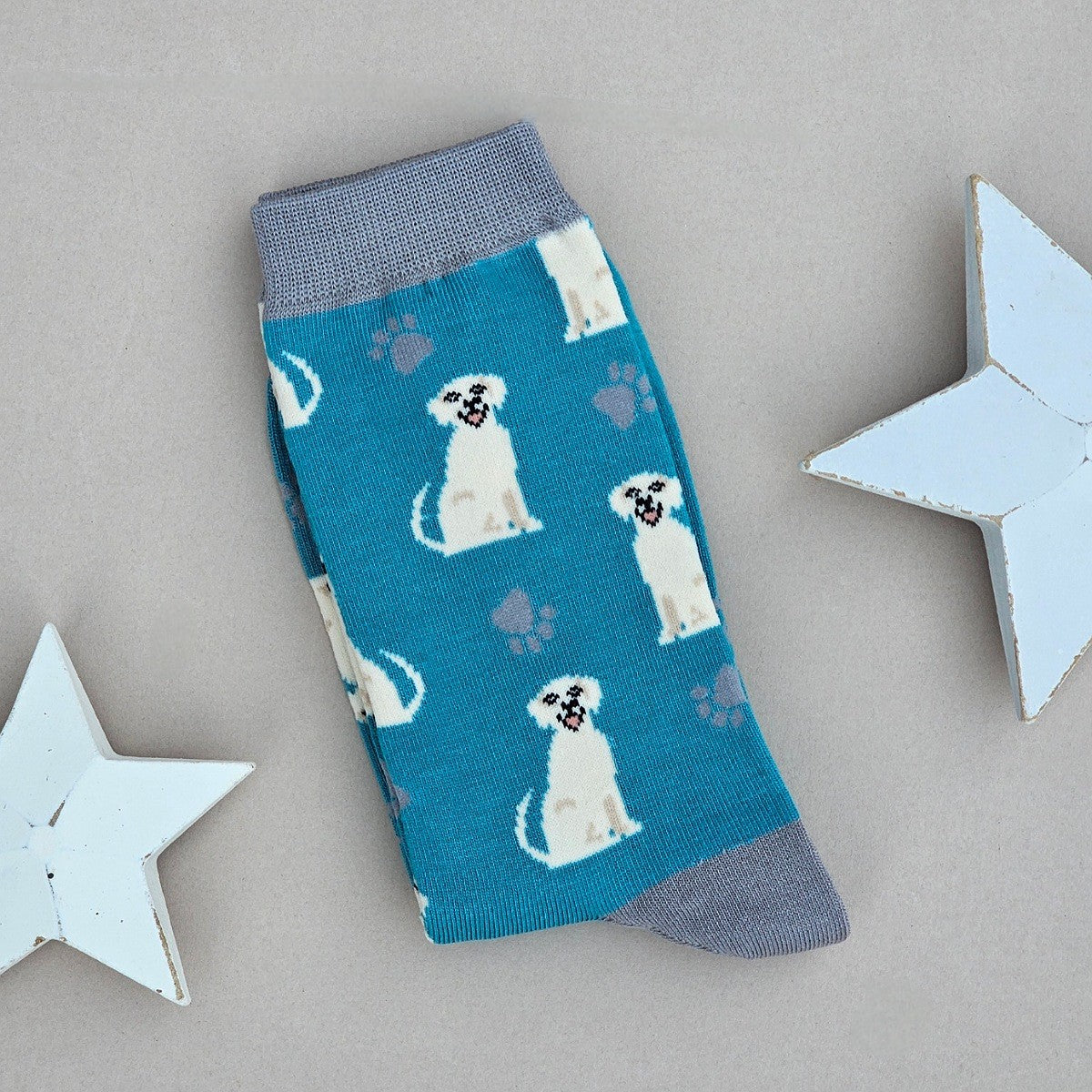 A single blue sock with an all-over  pattern of smiling labradors, with grey printed paws.  Silver grey ribbed cuffs and heels, styled on a light grey surface with white star props.

