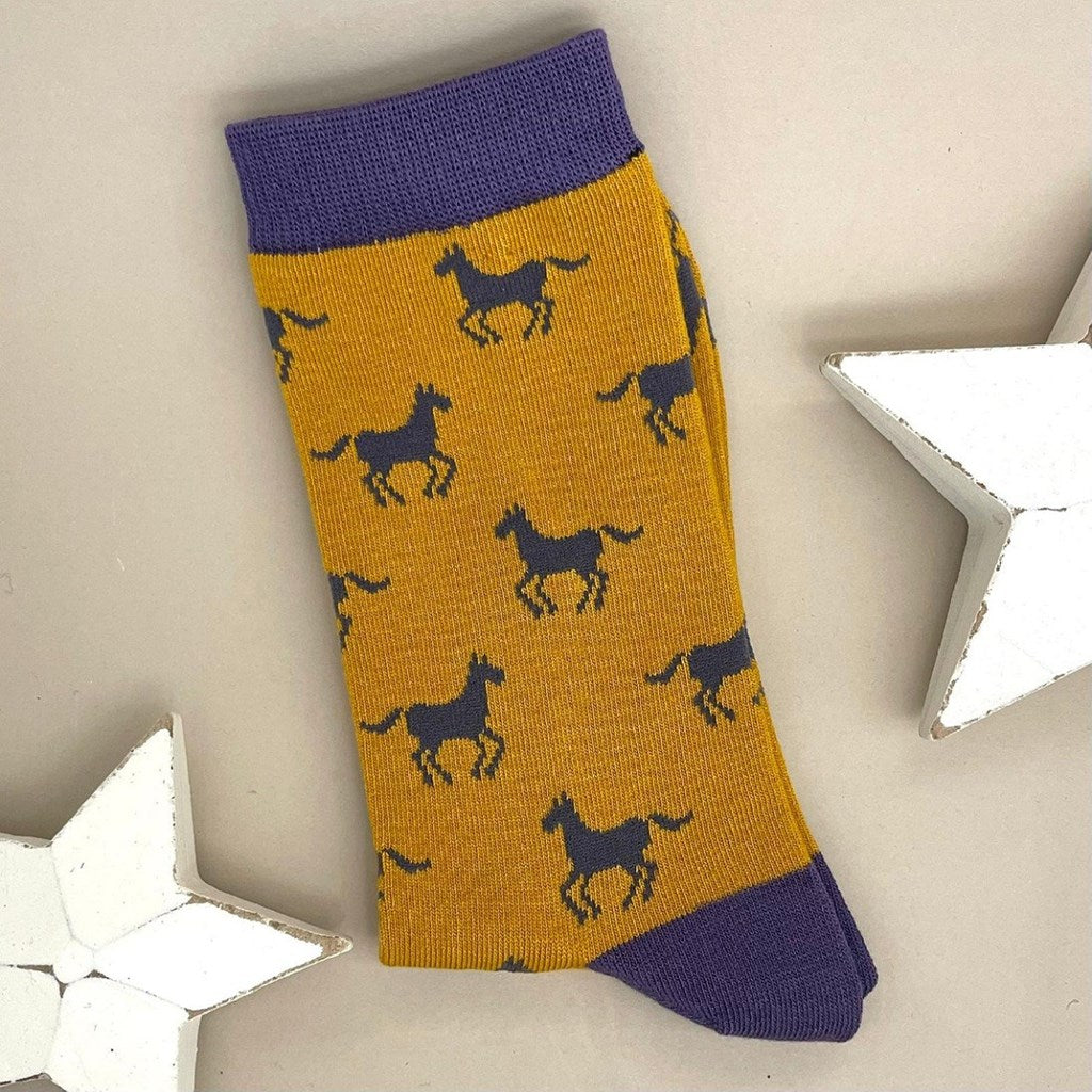 A single mustard sock with grey horse silhouettes.  Purple cuff, heel, and toe, laid flat on a light beige background with two white star decorations.
