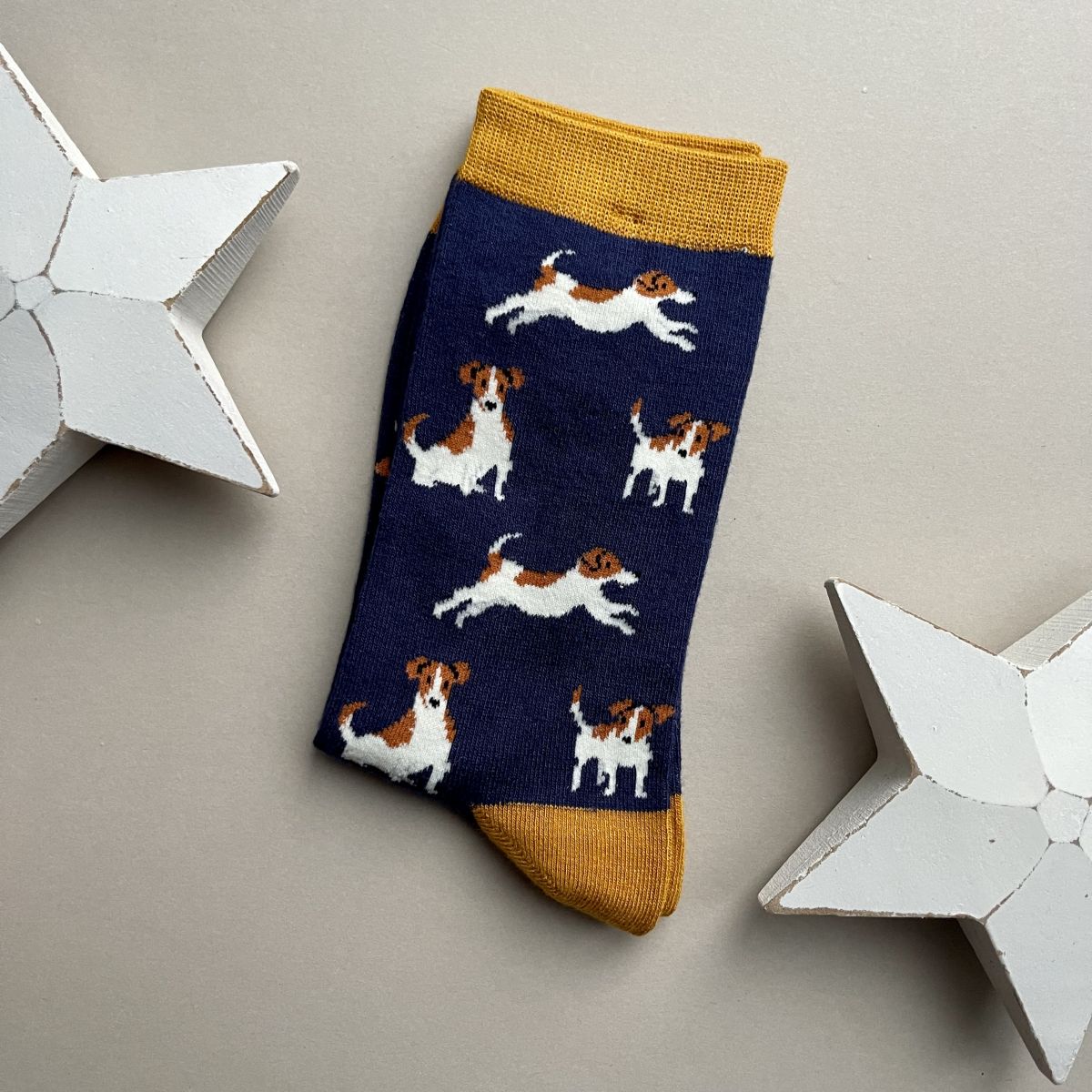 A single navy sock with an all-over pattern of various white and brown jack Russell dogs.  Mustard cuff and heel, displayed on a neutral beige background with white star-shaped props.