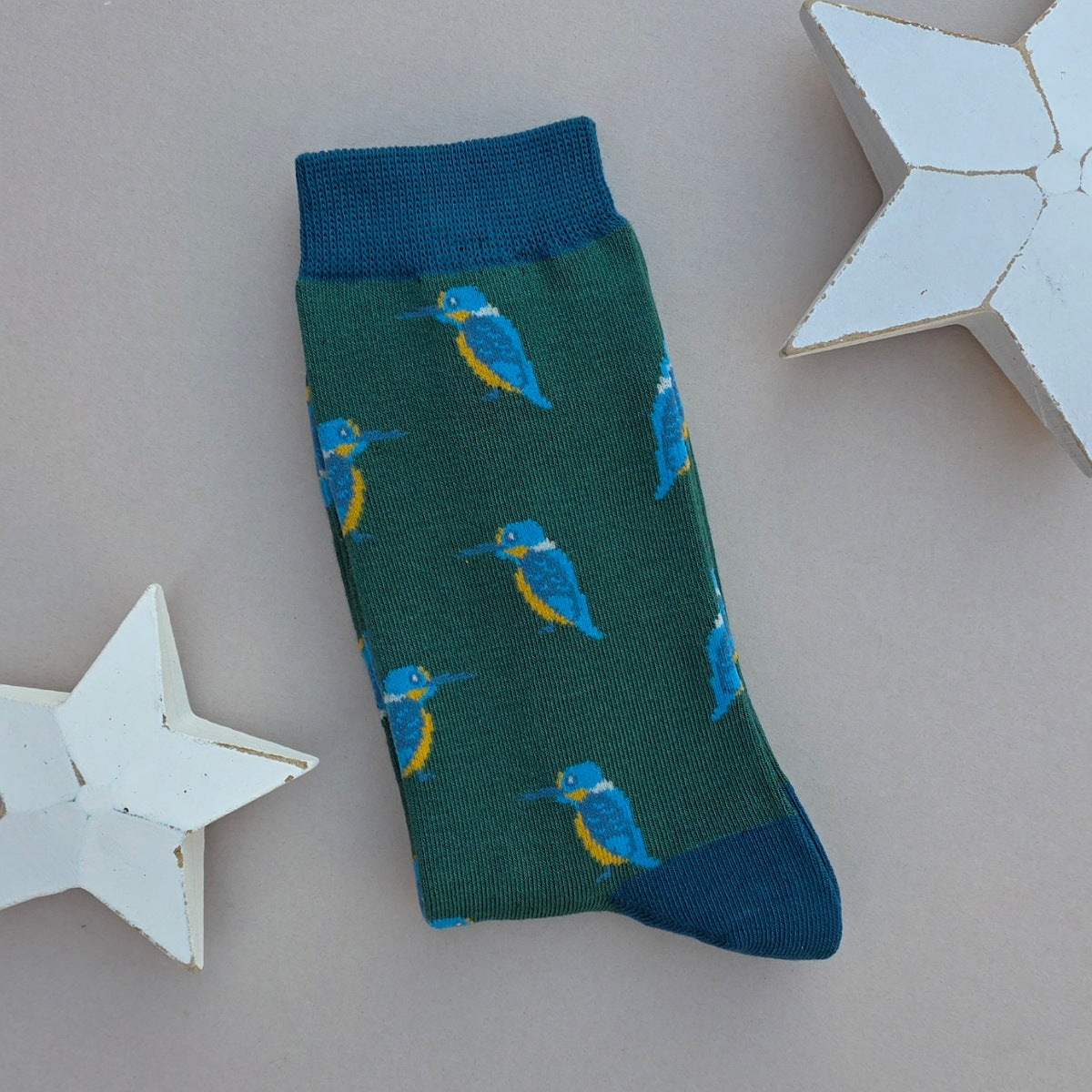 A single green sock with a repeating pattern of blue and yellow kingfisher birds, featuring a teal cuff and toe, displayed on a neutral beige background with white star-shaped props.
