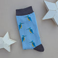 A single light blue sock with a repeating pattern of blue and yellow kingfisher birds, featuring a grey cuff and toe, displayed on a neutral beige background with white star-shaped props.