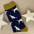 A single navy blue sock with an all-over  pattern of light coloured labradors.  Yellow ribbed cuffs and heels, styled on a light grey surface with white star props.
