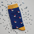 A single navy sock with an all-over  pattern of little robins.  Mustard ribbed cuffs and heels.  Styled on a light grey surface with scattered silver stars props.