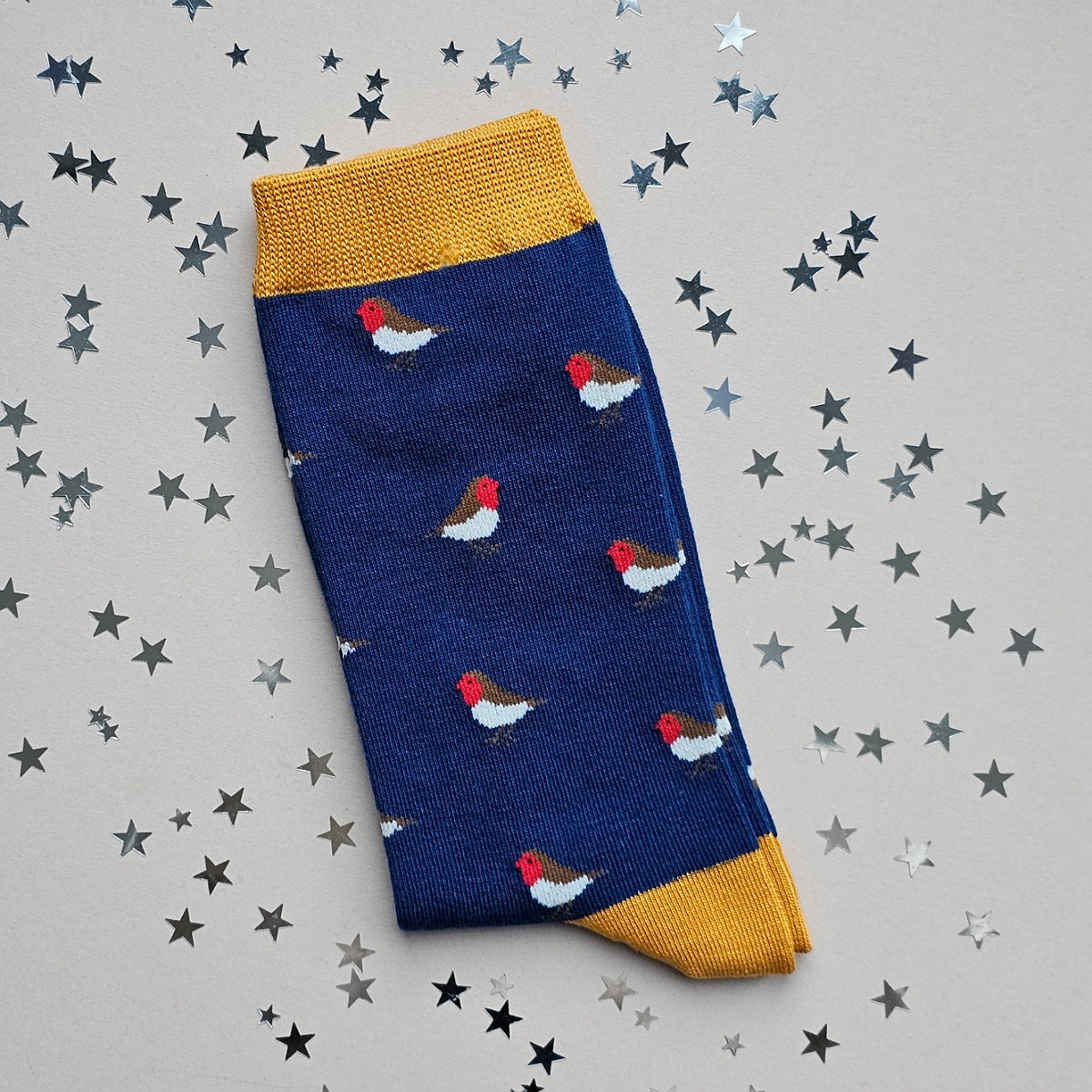 A single navy sock with an all-over  pattern of little robins.  Mustard ribbed cuffs and heels.  Styled on a light grey surface with scattered silver stars props.