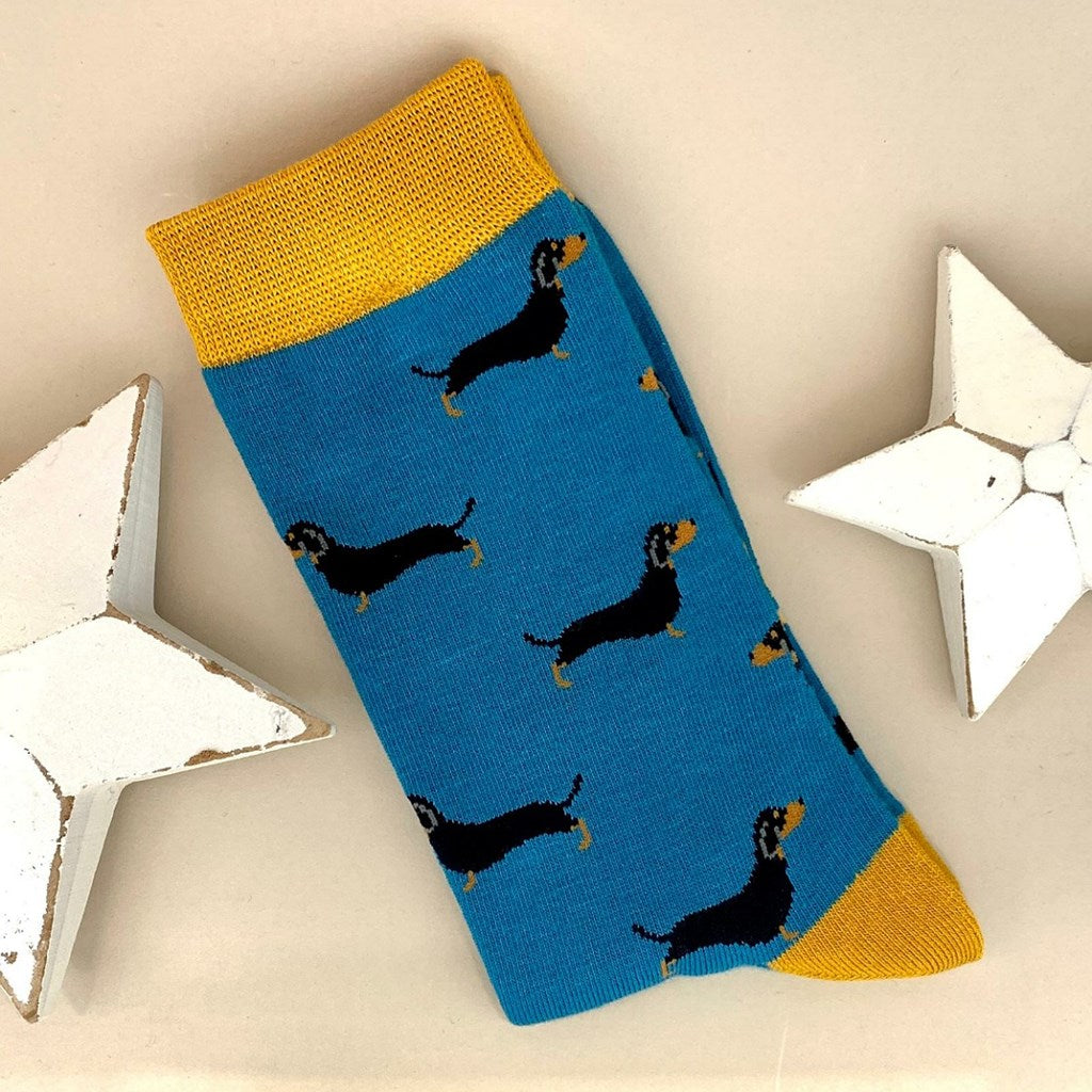 A single teal sock with little black sausage dog silhouettes and yellow cuff, heel, and toe, laid flat on a light beige background with two white star decorations. 