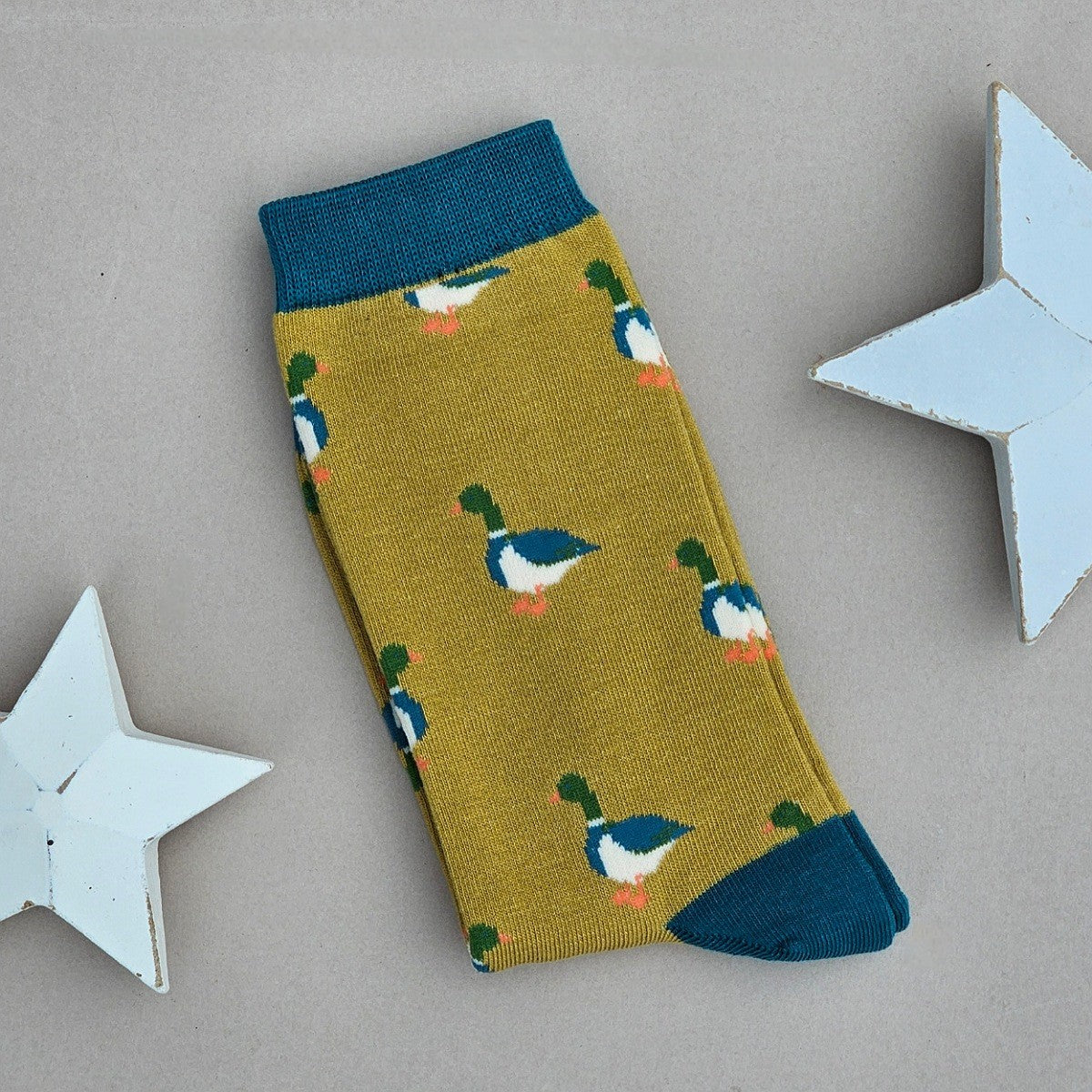 A single mustard sock with various mallard duck silhouettes and teal yellow cuff, heel, and toe, laid flat on a light beige background with two white star decorations.