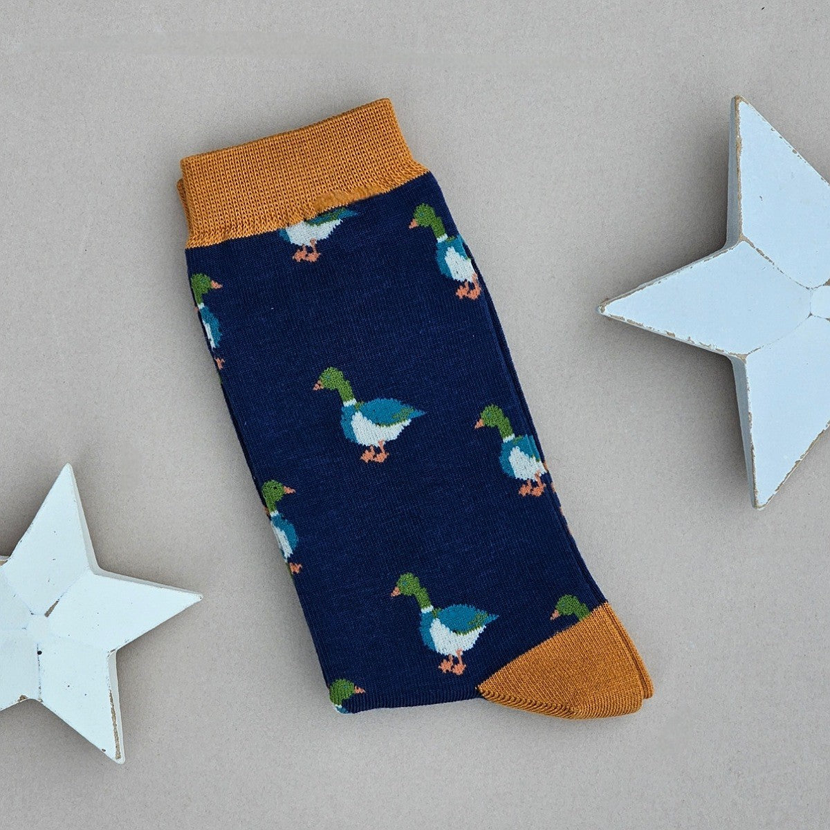 A single navy sock with various mallard duck silhouettes and mustard yellow cuff, heel, and toe, laid flat on a light beige background with two white star decorations.