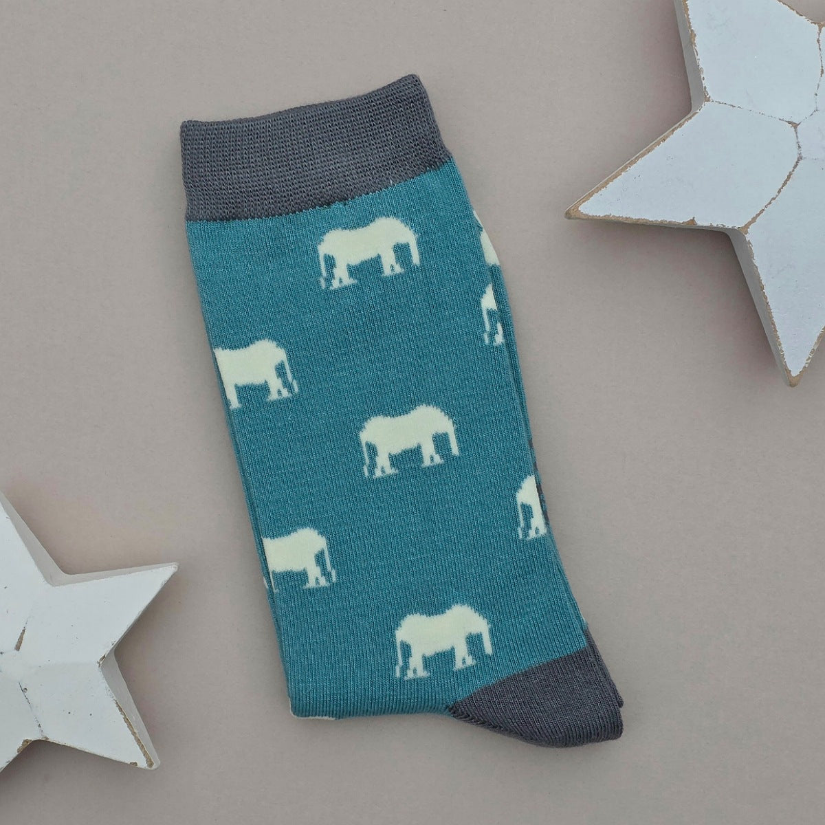 A single teal sock with light coloured elephant silhouettes and grey yellow cuff, heel, and toe, laid flat on a light beige background with two white star decorations.