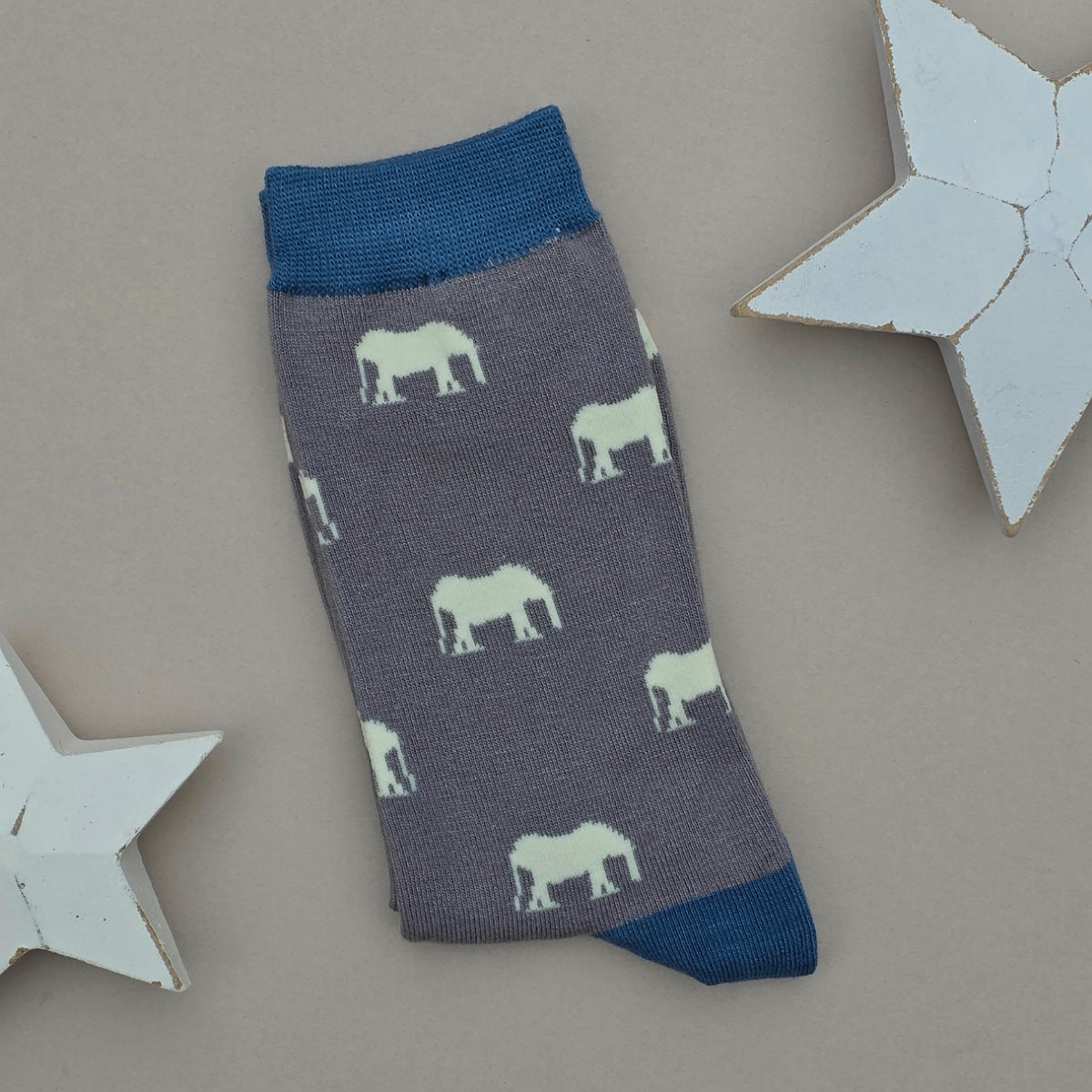 A single grey sock with light grey elephant silhouettes and teal cuff, heel, and toe, laid flat on a light beige background with two white star decorations.