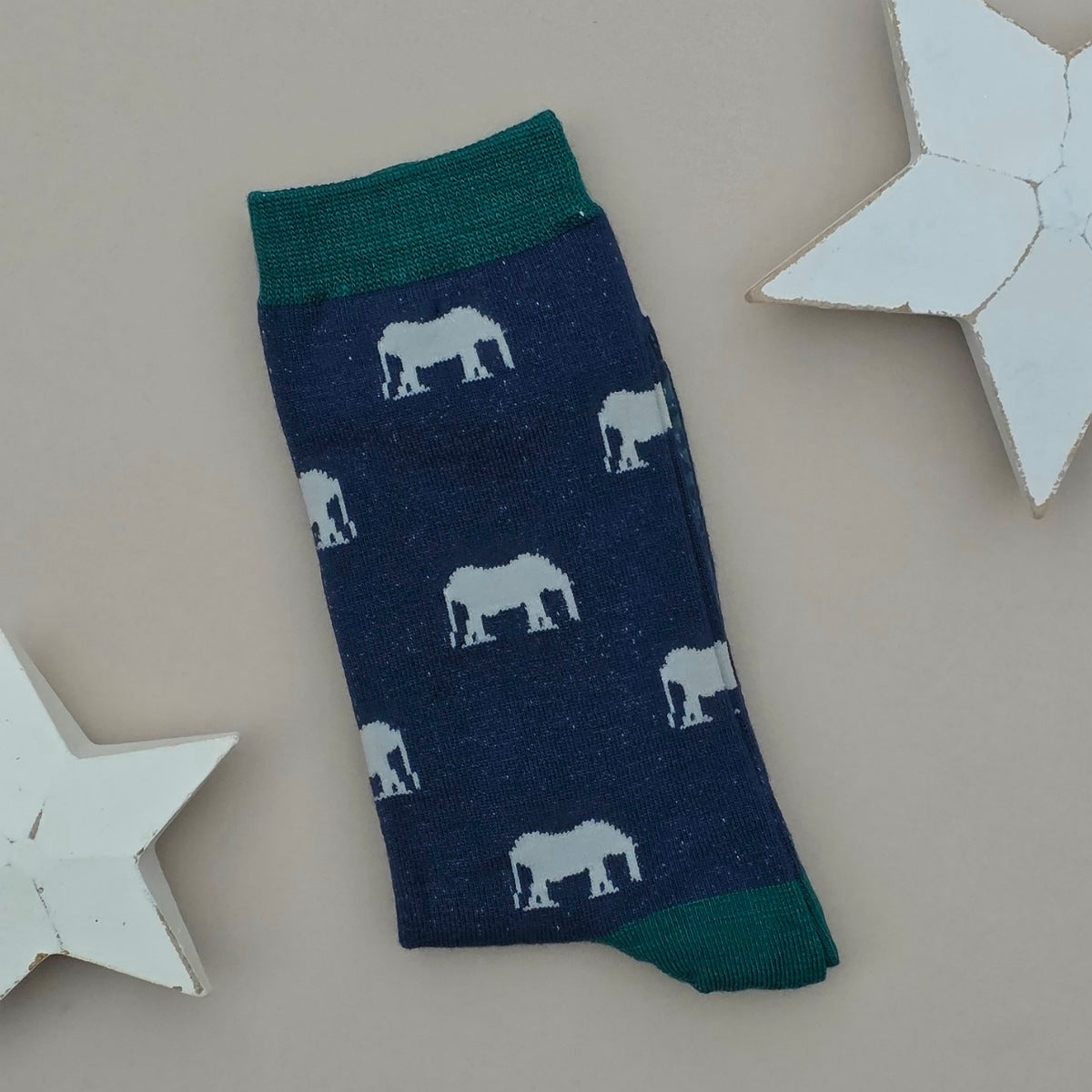 A single navy sock with grey elephant silhouettes and teal cuff, heel, and toe, laid flat on a light beige background with two white star decorations.