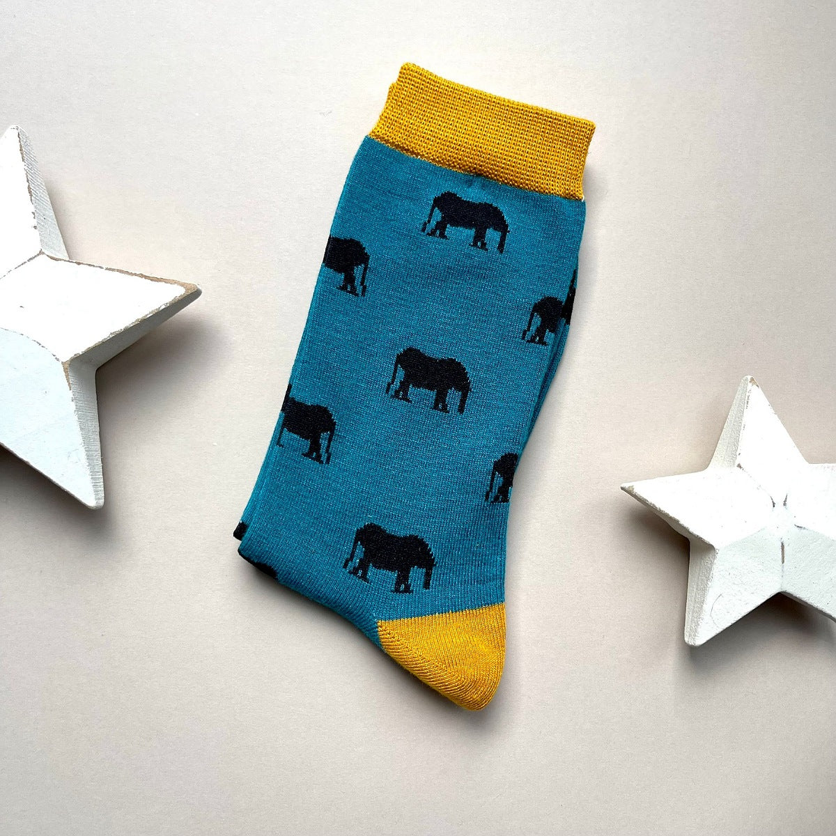 A single blue sock with black elephant silhouettes and mustard yellow cuff, heel, and toe, laid flat on a light beige background with two white star decorations.