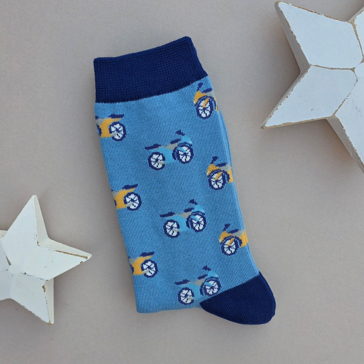 A single blue sock with an all-over  pattern of blue and yellow motor bikes, navy blue ribbed cuffs and heels, styled on a light grey surface with white star props.