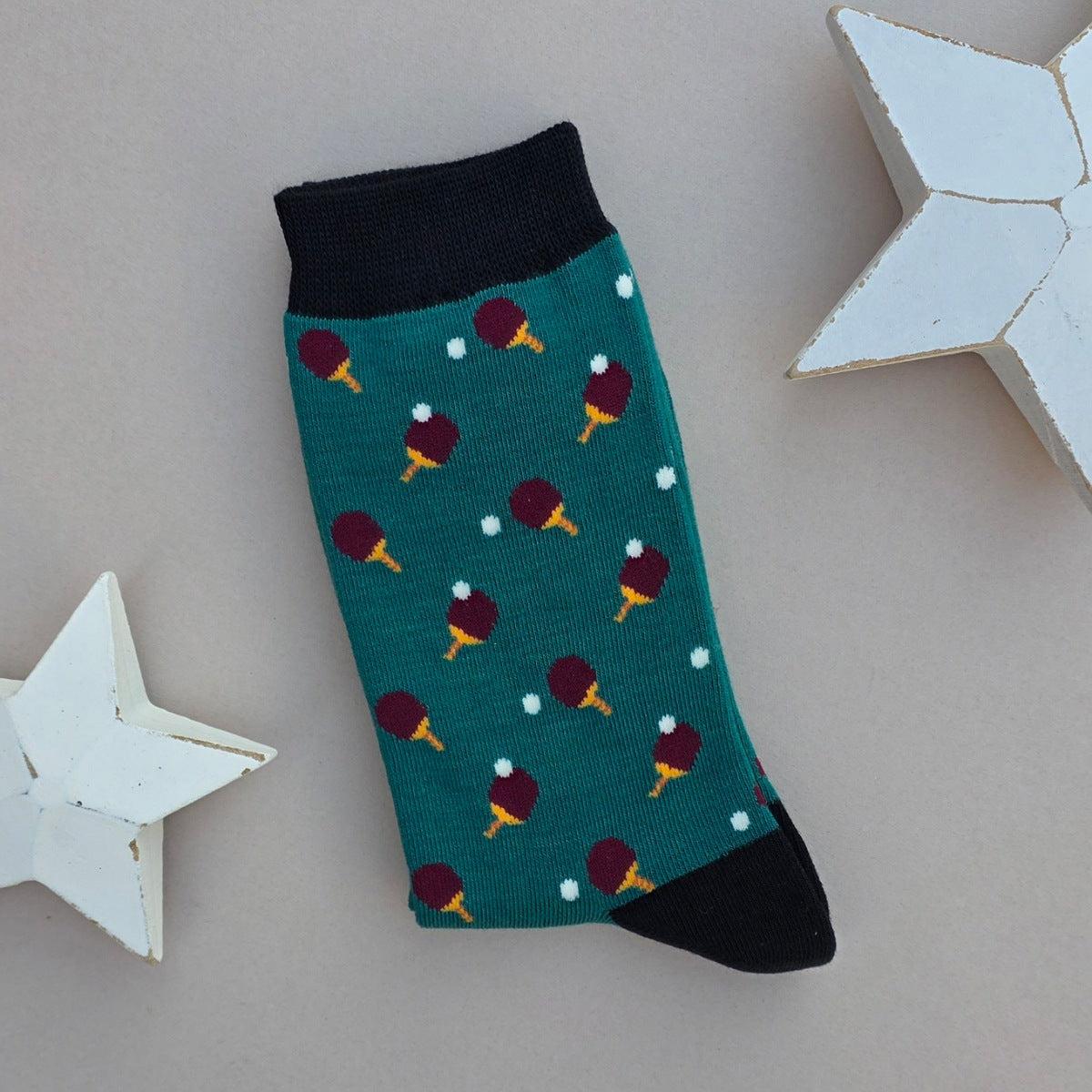 A single green sock with an all-over  pattern of ping pong bat and balls.  Black ribbed cuffs and heels, styled on a light grey surface with white star props.