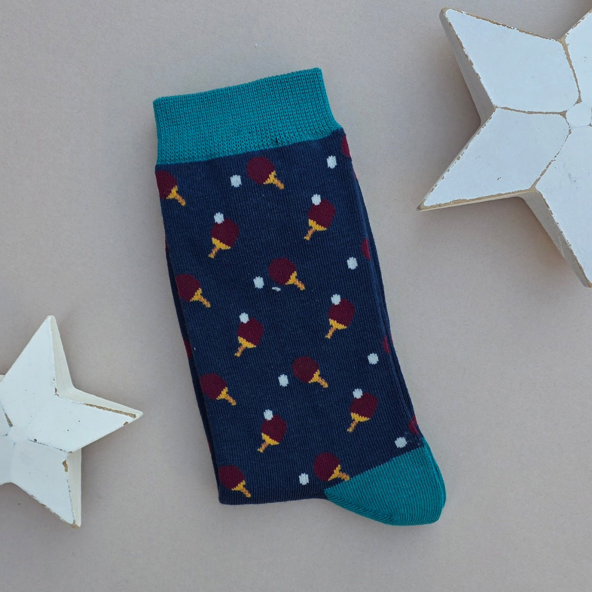 A single navy blue sock with an all-over  pattern of ping pong bat and balls.  Teal  ribbed cuffs and heels, styled on a light grey surface with white star props
