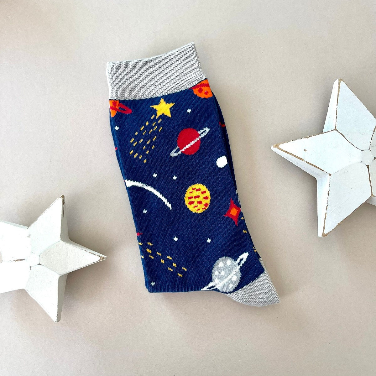 A single navy blue sock with a colourful space-themed print of planets, stars, and rockets with light grey cuff, heal and toe.  Styled on a light grey background with two white star-shaped props.