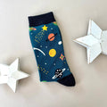 A single teal sock with a colourful space-themed print of planets, stars, and rockets with black cuff, heal and toe.  Styled on a light grey background with two white star-shaped props.